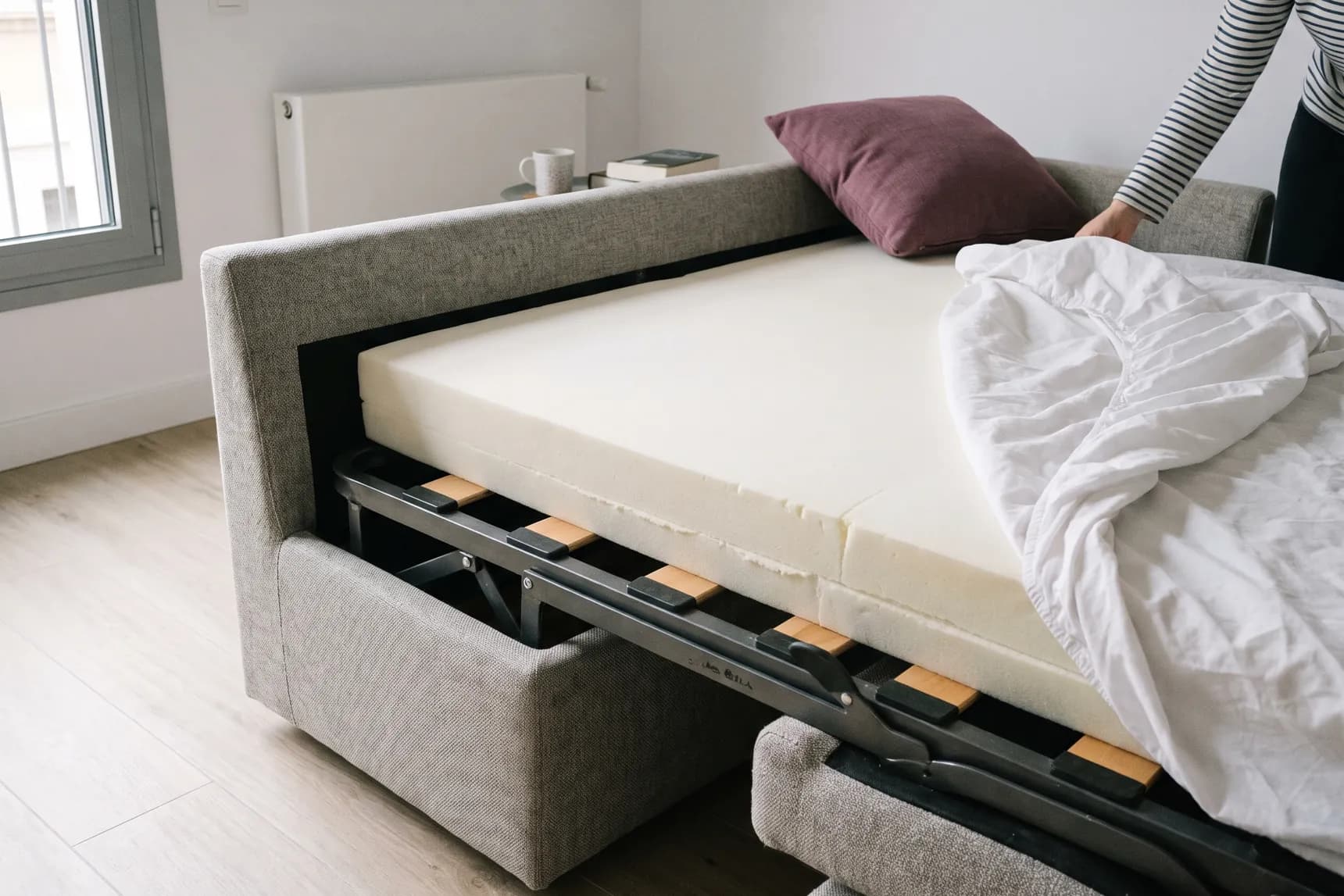Colchón grueso de espuma sobre el somier de un sofá cama italiano, con la sábana a medio poner (imagen generada con IA)