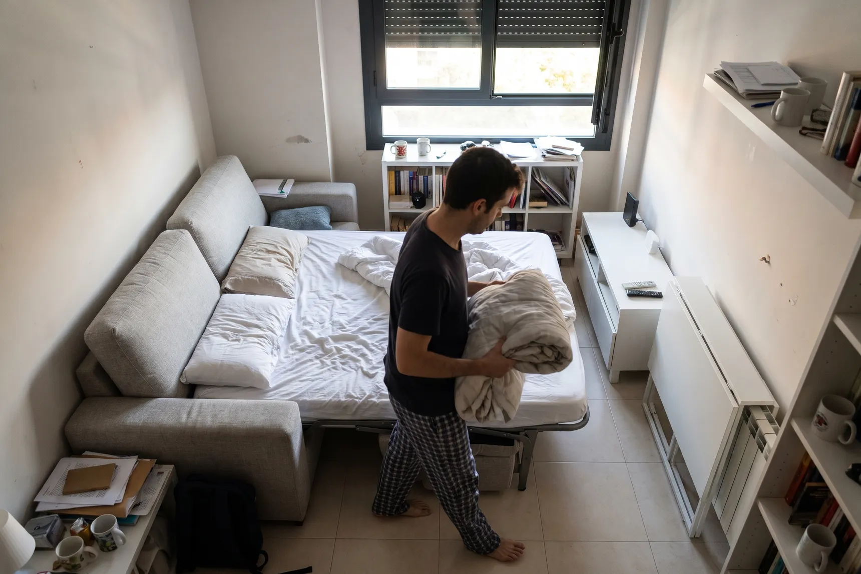 Salón pequeño con un sofá cama abierto que ocupa casi todo el suelo hasta el mueble de la tele; una persona pasa de lado con el edredón (imagen generada con IA)